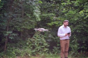 In focus, an aerial drone and in the background out of focus, a man in a hat who is flying the drone